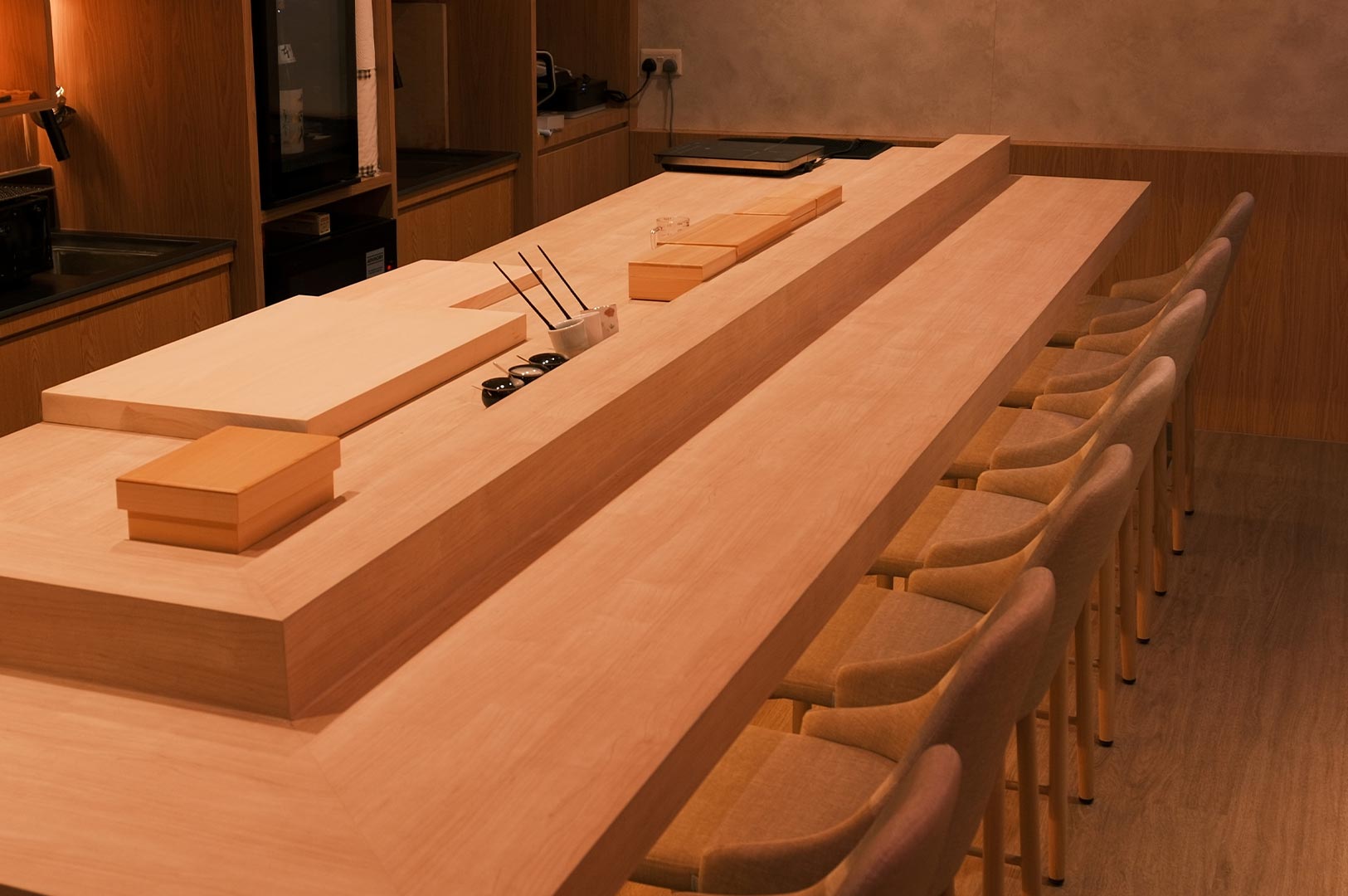 A wide-angle interior shot of a minimalist Japanese omakase restaurant featuring a long, light-wood sushi counter with high-back chairs and traditional wooden prep boxes under warm, ambient lighting.
