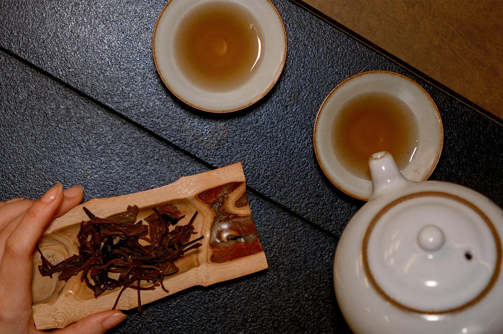 A top-down view of a person pouring amber-colored tea from a white ceramic teapot into a small cup, with dark dried oolong tea leaves presented on a carved bamboo tea scoop.