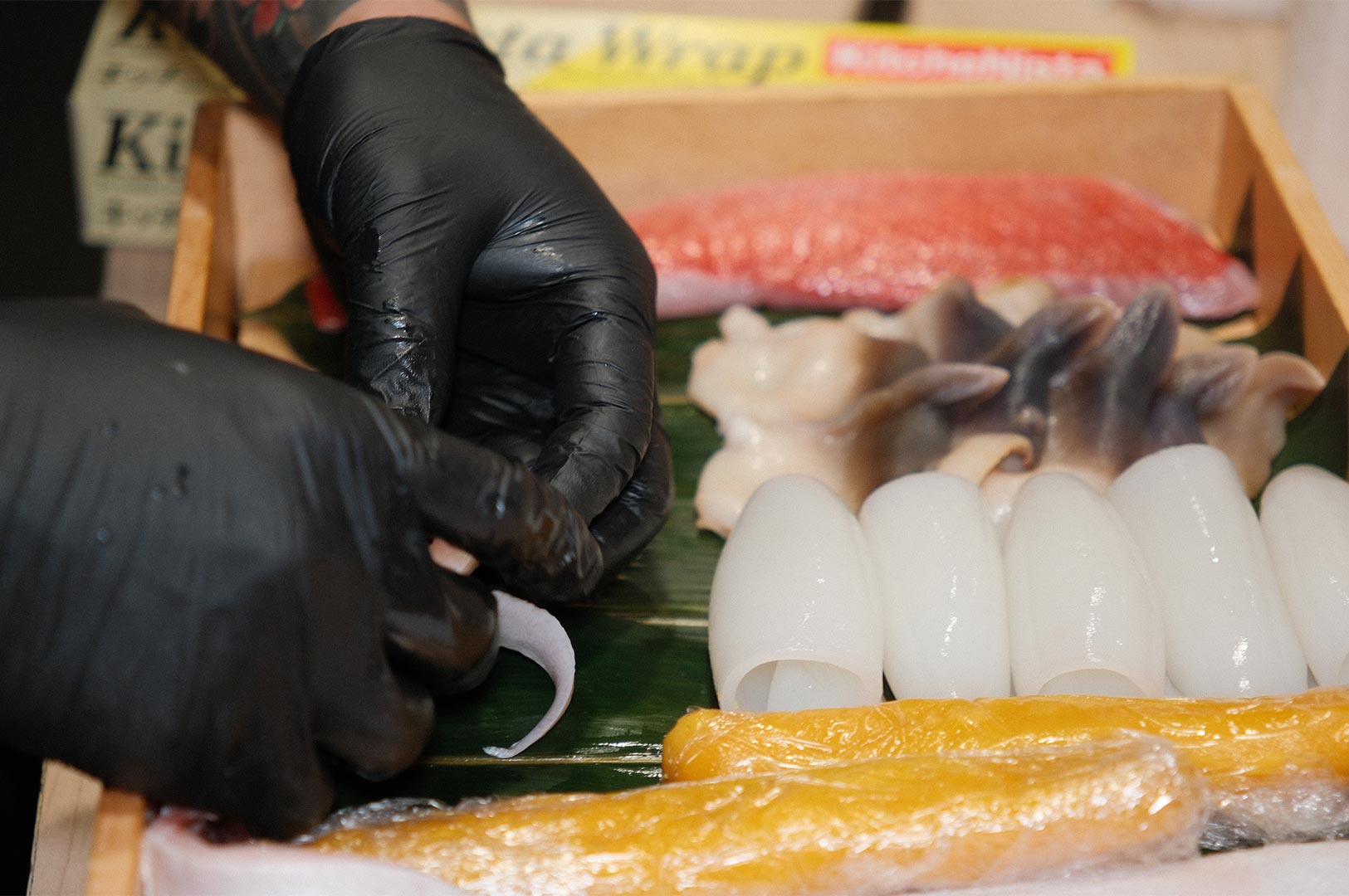 Close-up of a sushi chef's hands in black gloves carefully preparing fresh seafood, including squid, tuna, and shellfish, displayed on a traditional bamboo leaf inside a wooden box.