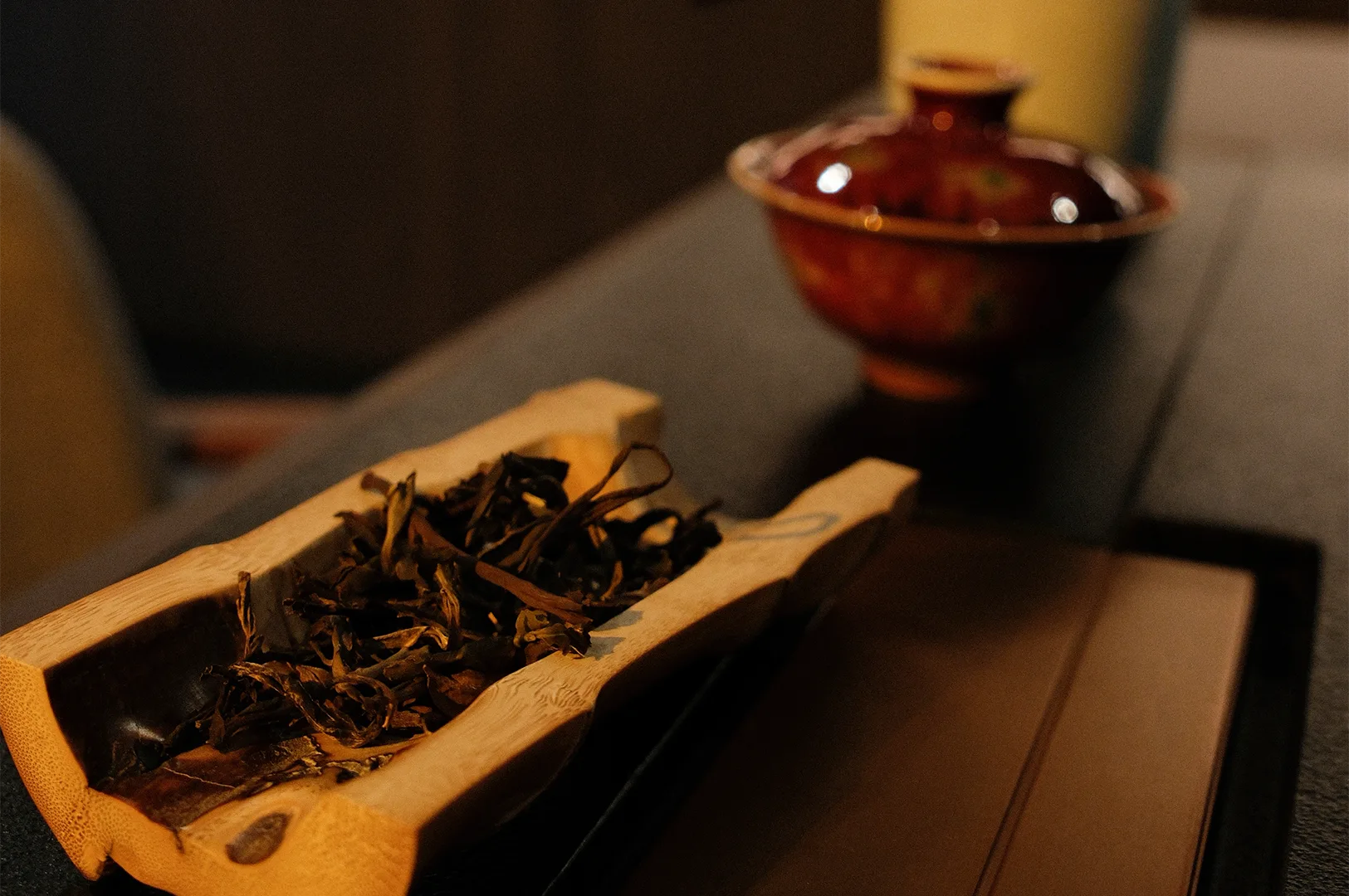 High-angle shot of dried loose leaf tea in a traditional bamboo tea holder (cha he) on a dark tea table with a blurred gaiwan in the background.
