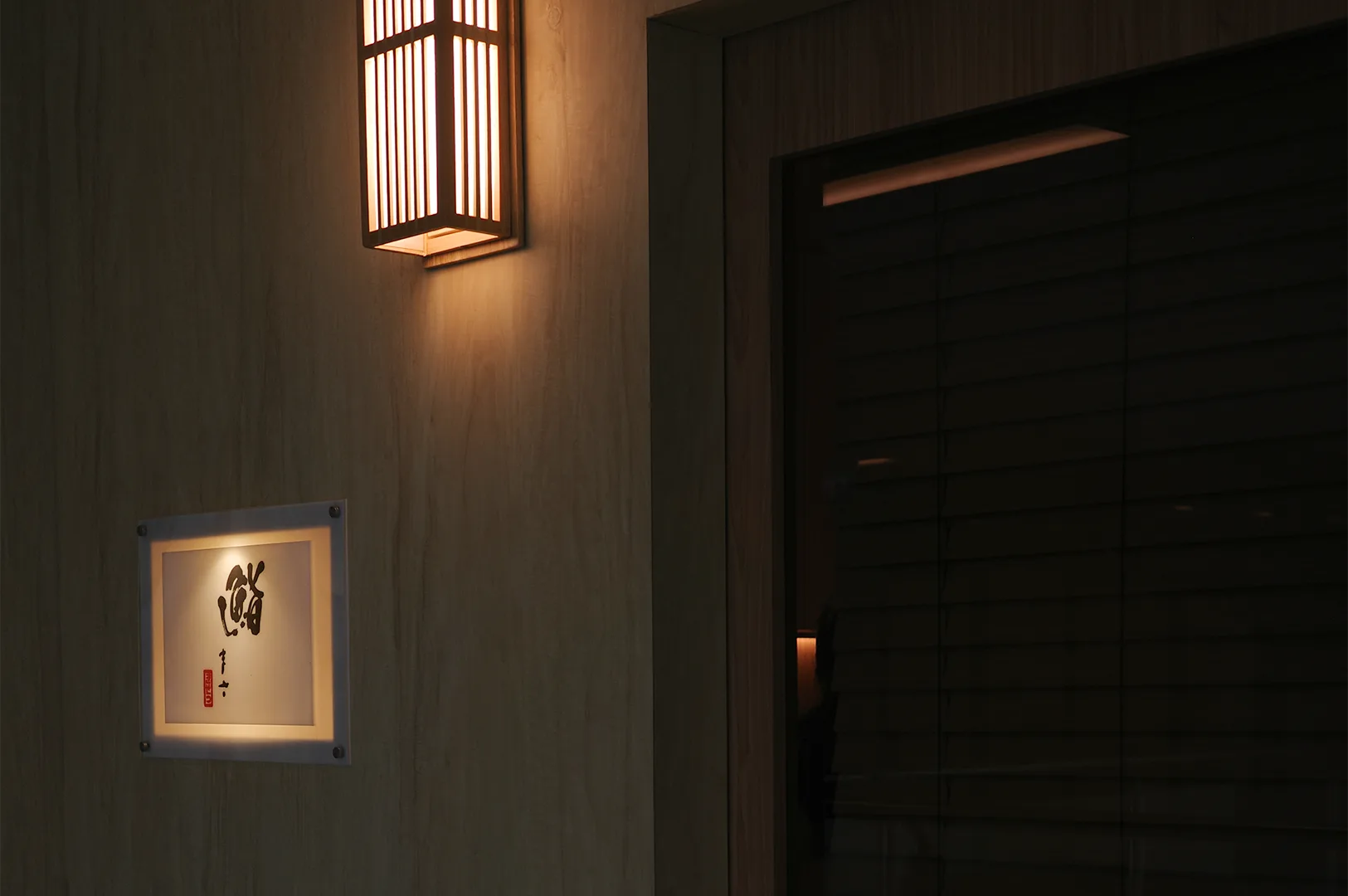 A minimalist wooden entrance of a Japanese sushi restaurant featuring a traditional warm lantern and a backlit calligraphy sign.