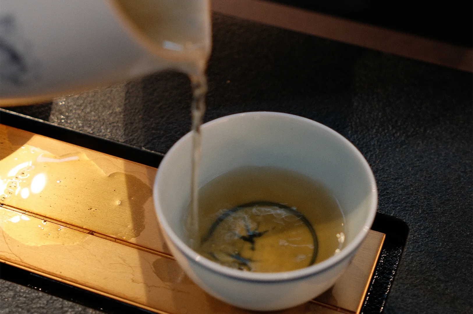 A close-up shot of light golden oolong tea being poured from a white ceramic pitcher into a matching tea bowl on a wooden tea tray.