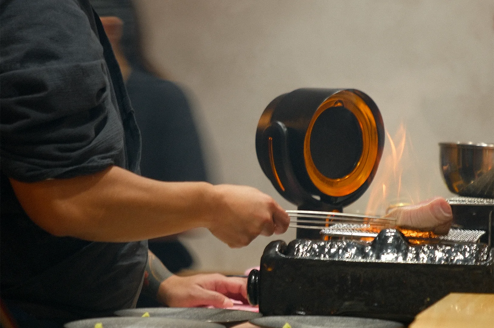 A Japanese sushi chef using a hand-held charcoal grill to sear a piece of fatty tuna (toro) over an open flame.