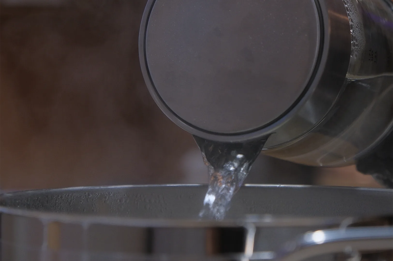 A close-up shot of clear boiling water being poured from a modern kettle into a stainless steel pot, creating steam.