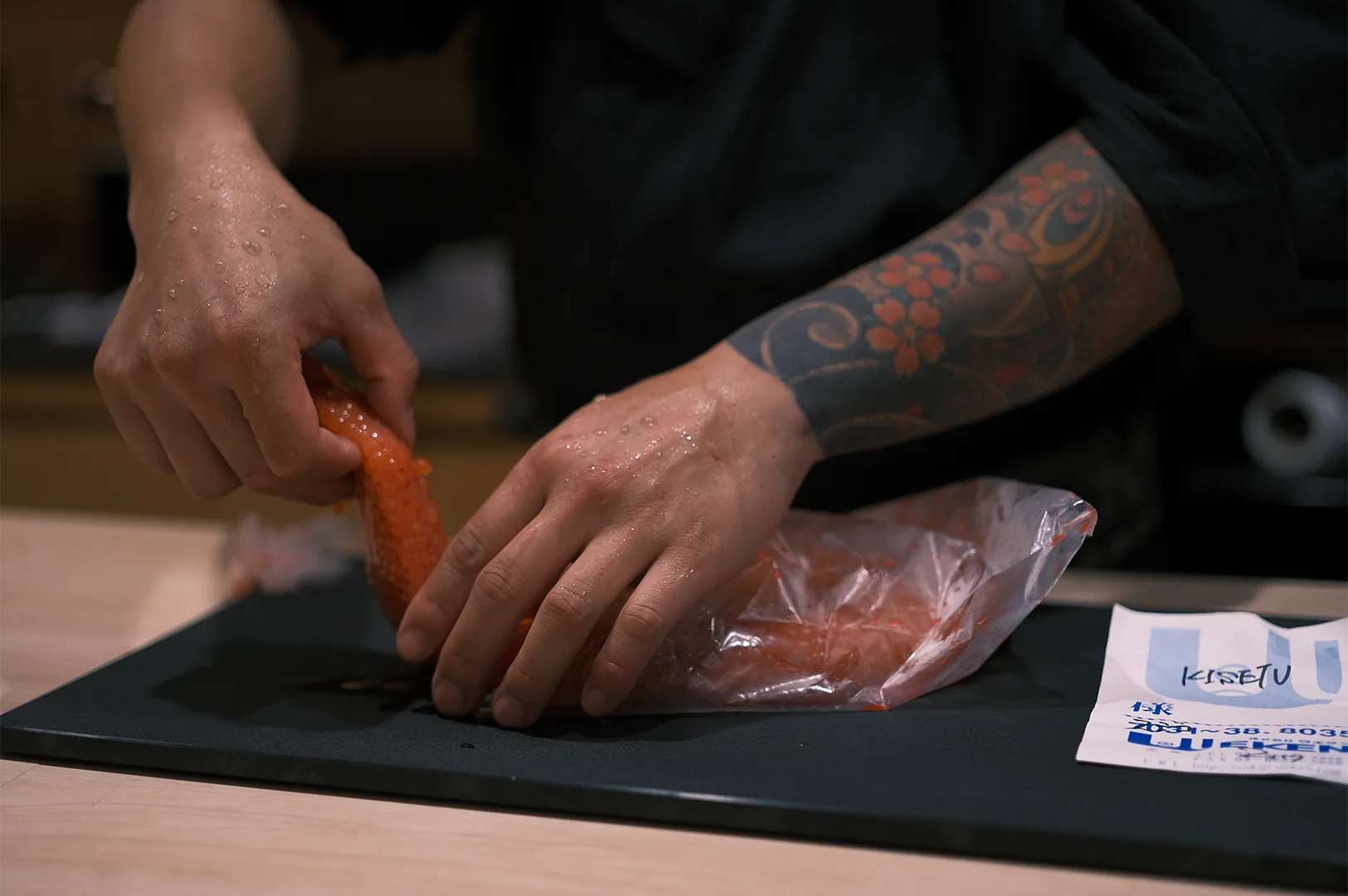 A professional sushi chef with a traditional forearm tattoo carefully handling a sac of fresh orange salmon roe (sujiko) on a black cutting board.