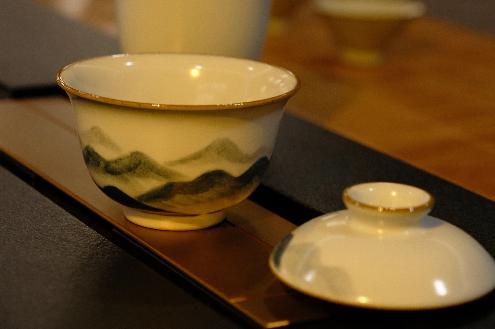 A traditional Chinese Gaiwan tea cup featuring a hand-painted green mountain landscape design with its lid resting beside it on a dark textured surface.