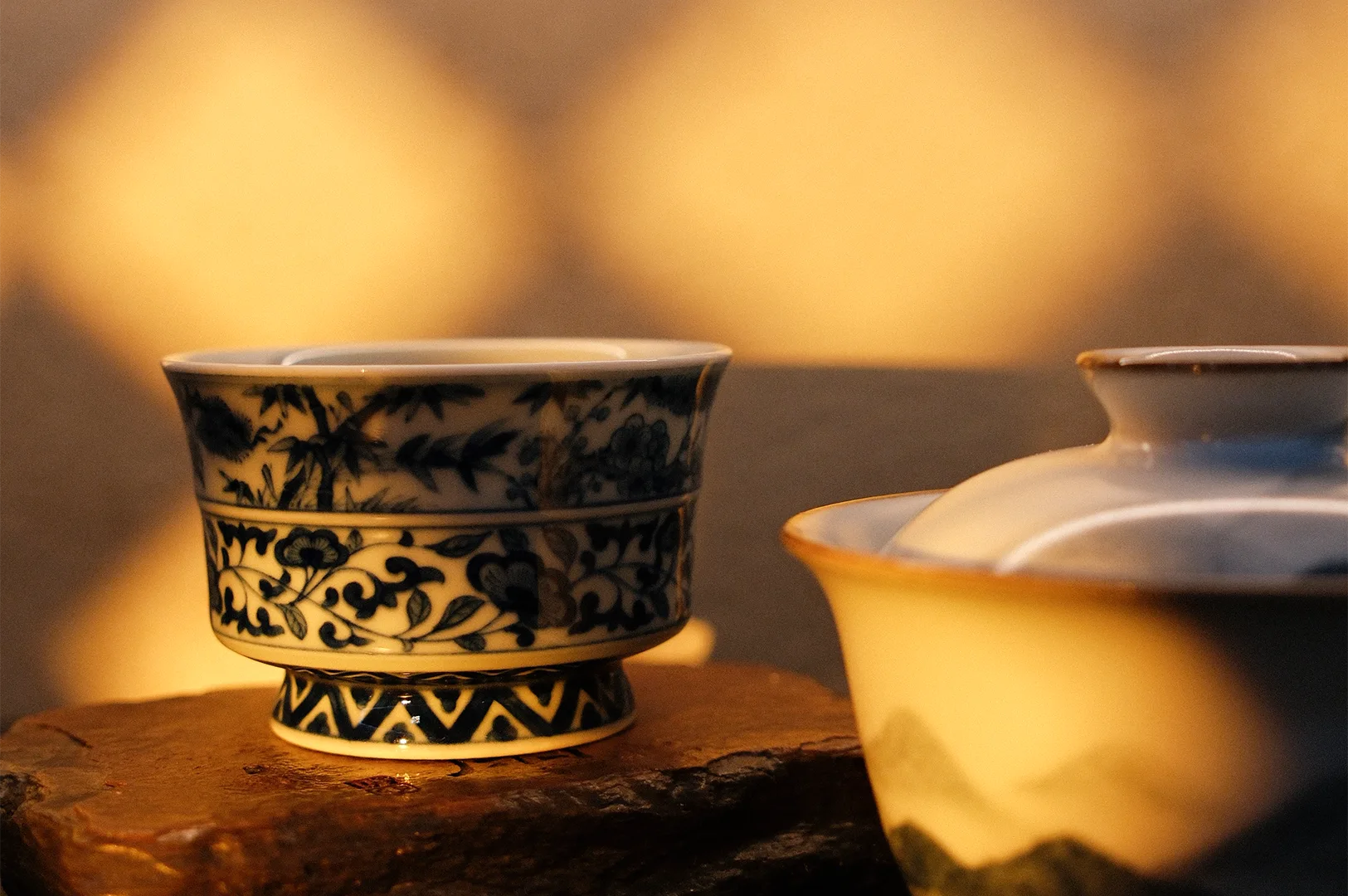 A close-up of a traditional Chinese blue and white porcelain tea cup and a lidded gaiwan resting on a rustic wooden base under warm, atmospheric lighting.