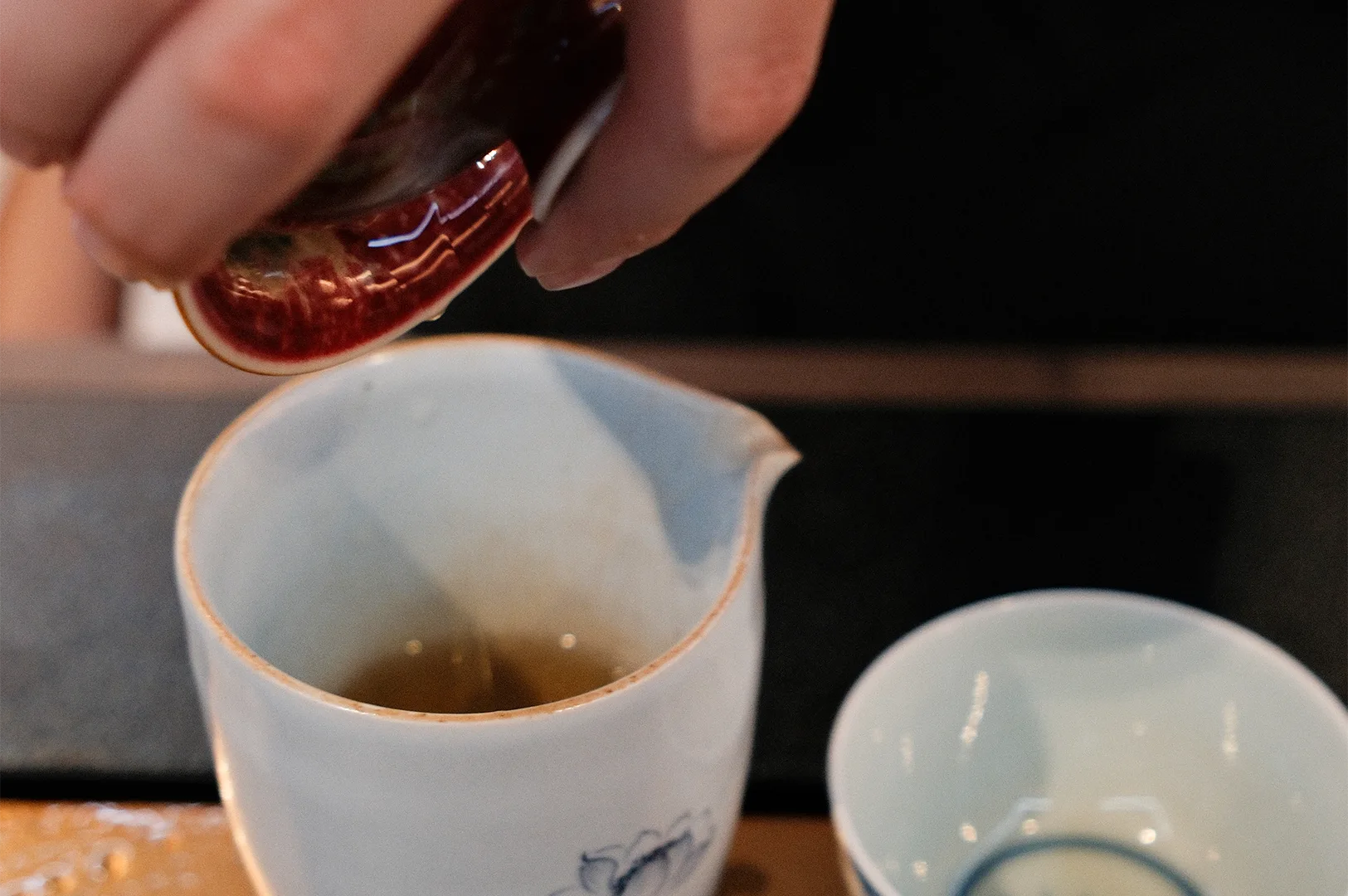 A person pouring freshly brewed tea from a ceramic vessel into a white porcelain fairness cup (Gongdao Bei) during a traditional tea ceremony.