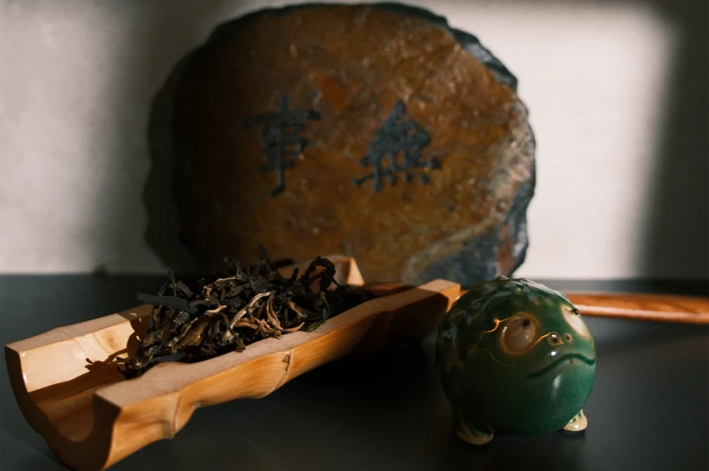 Premium loose-leaf tea leaves on a carved bamboo scoop, featuring a green ceramic frog tea pet and a stone plaque with Chinese calligraphy in the background.