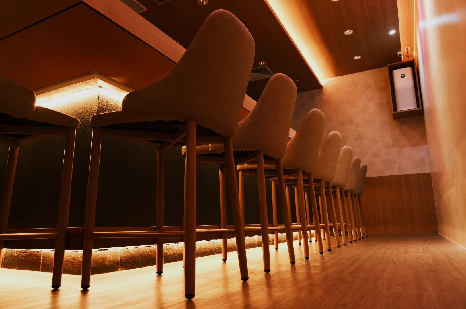 Modern bar interior with a low-angle view of a row of beige upholstered barstools against a warm, softly lit wooden counter and floor. Cozy ambiance.