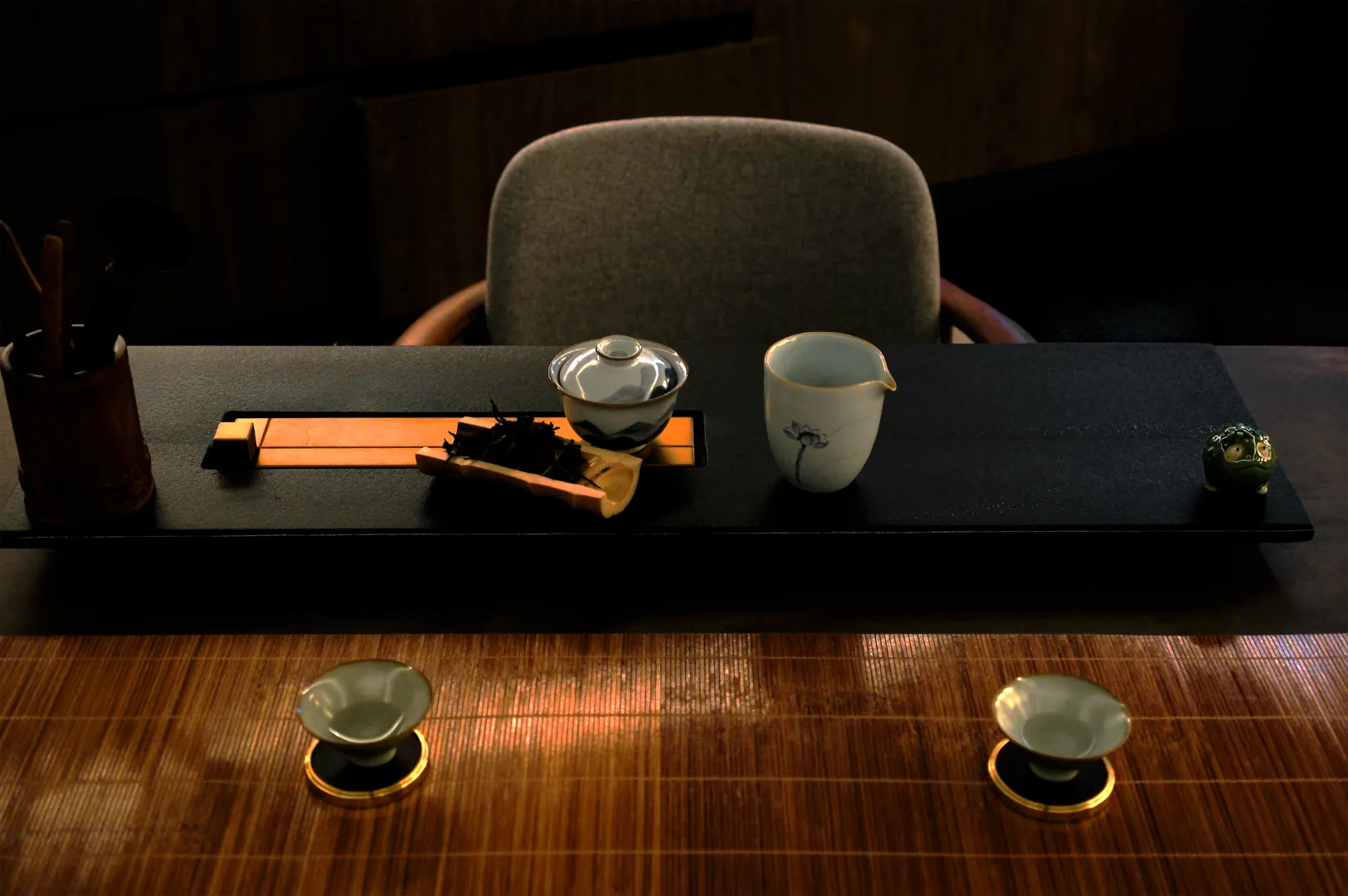 A moody tea setup with a black tray holding a covered teacup, pitcher, and tea leaves. Two small teacups are on a wooden table, evoking tranquility.