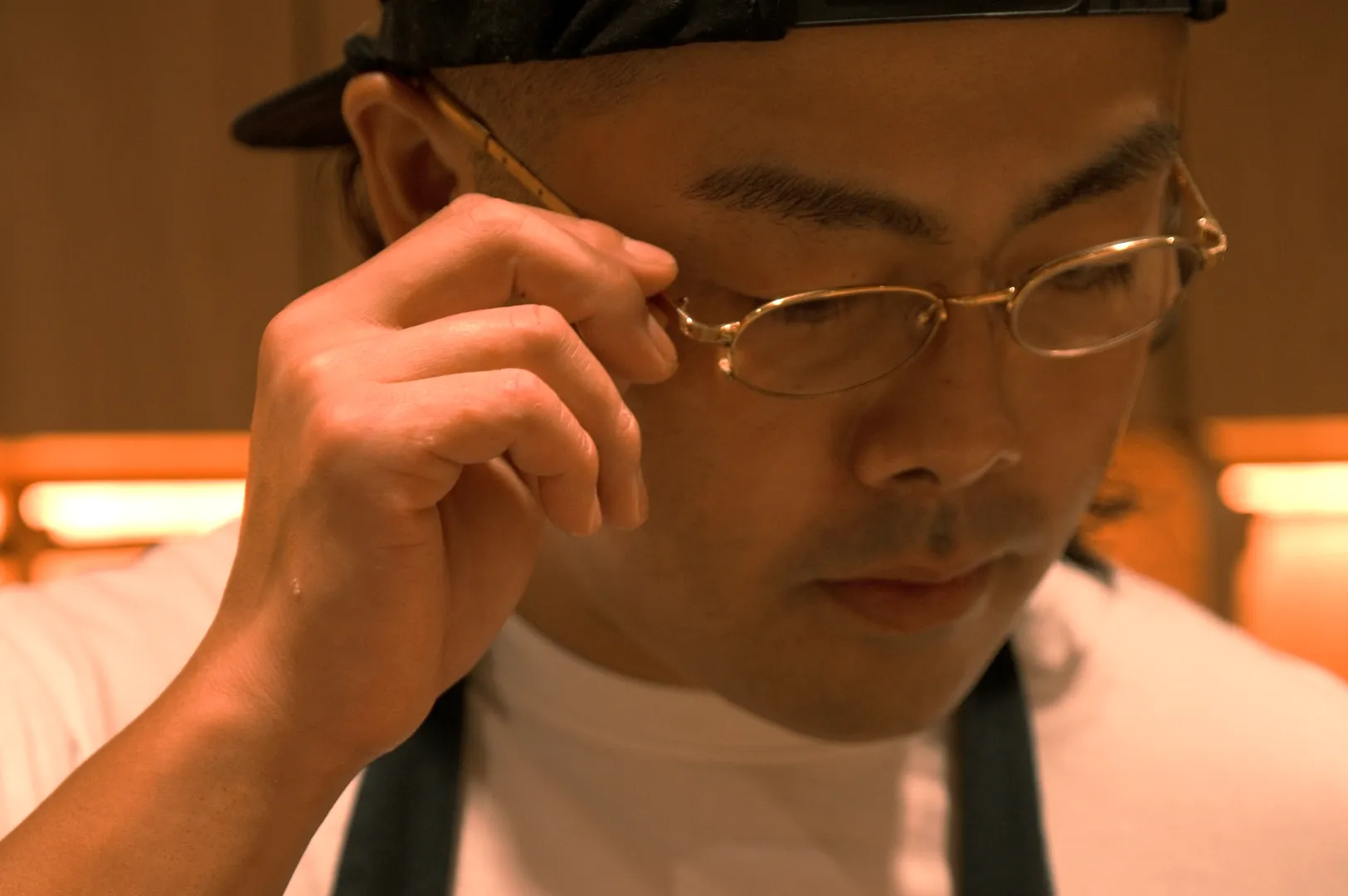 Chef Masa in a white shirt and black apron adjusts their glasses, exuding focus and concentration in a warmly lit kitchen setting.