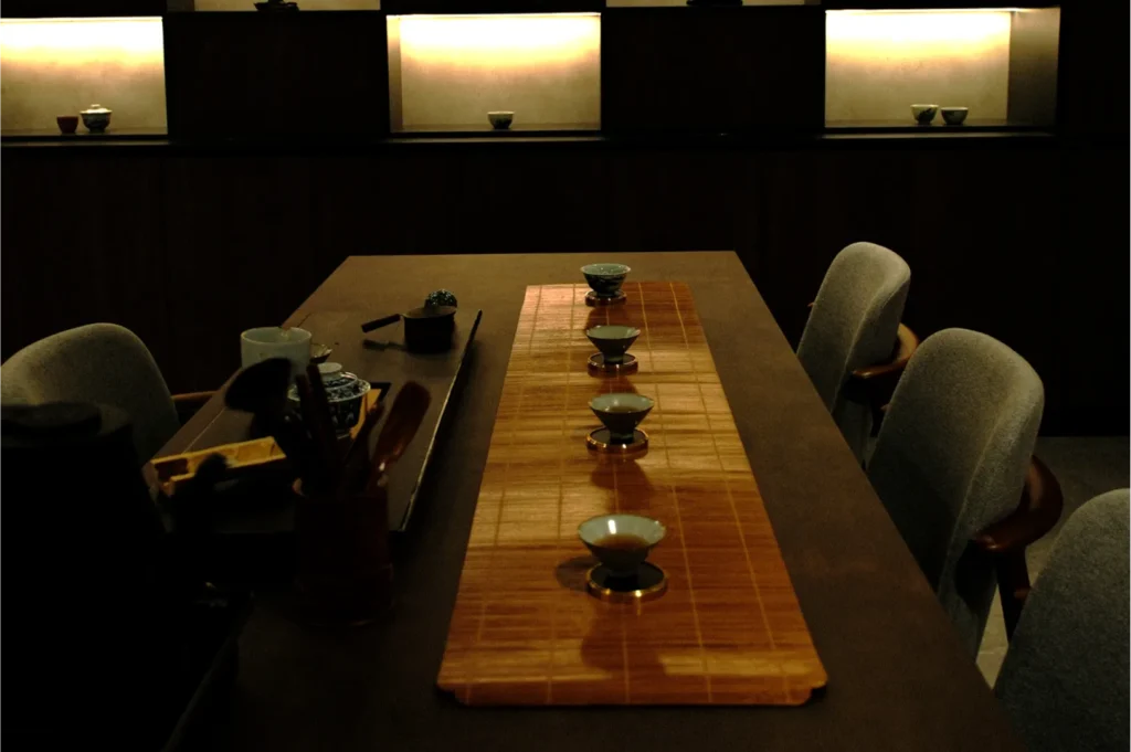 Dimly lit table set for tea with a bamboo mat and five tea bowls. Soft, backlit shelves in the background create an intimate, serene atmosphere.