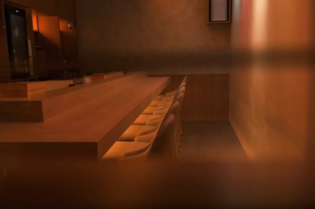 Dimly lit, cozy wooden bar interior with soft lighting, empty beige stools lined up neatly along the counter, creating a warm and inviting atmosphere.