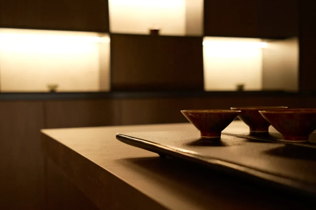 Minimalist interior with soft lighting features shelves in the background and a sleek table in the foreground. Three small ceramic bowls rest on a tray, evoking a serene and elegant atmosphere.