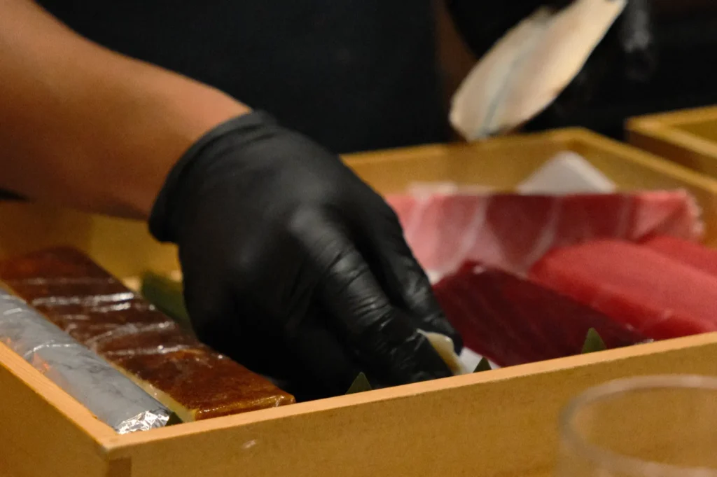 Chef Masa wearing a black glove places sushi pieces in a wooden box. The sushi includes various types of raw fish, creating an artisanal and fresh tone.