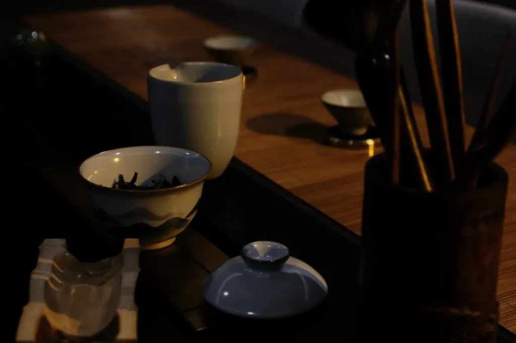 Dimly lit tea ceremony setup on a bamboo mat, featuring ceramic bowls, tea leaves, a wooden utensil holder, and a serene, traditional ambiance.