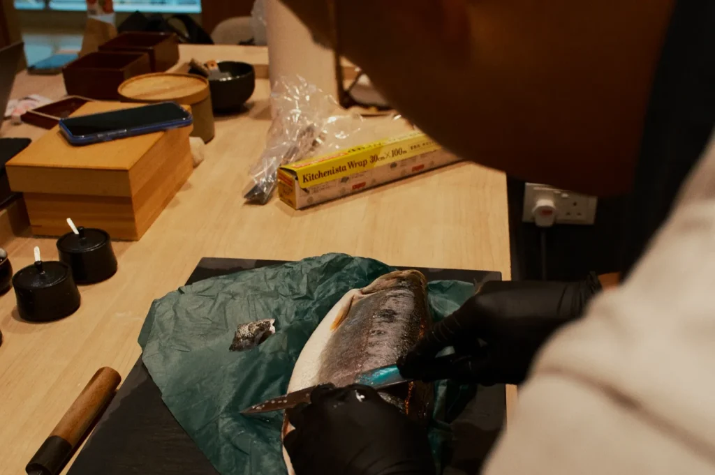 A person in black gloves fillets a large fish on a wooden table, surrounded by kitchen items and utensils, creating a focused and skilled atmosphere.