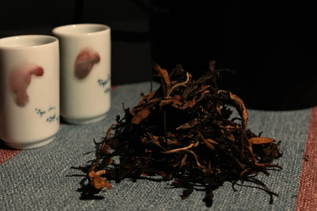 Loose tea leaves are piled on a textured blue surface, with two white ceramic cups featuring subtle red patterns in the background, creating a cozy, warm ambiance.