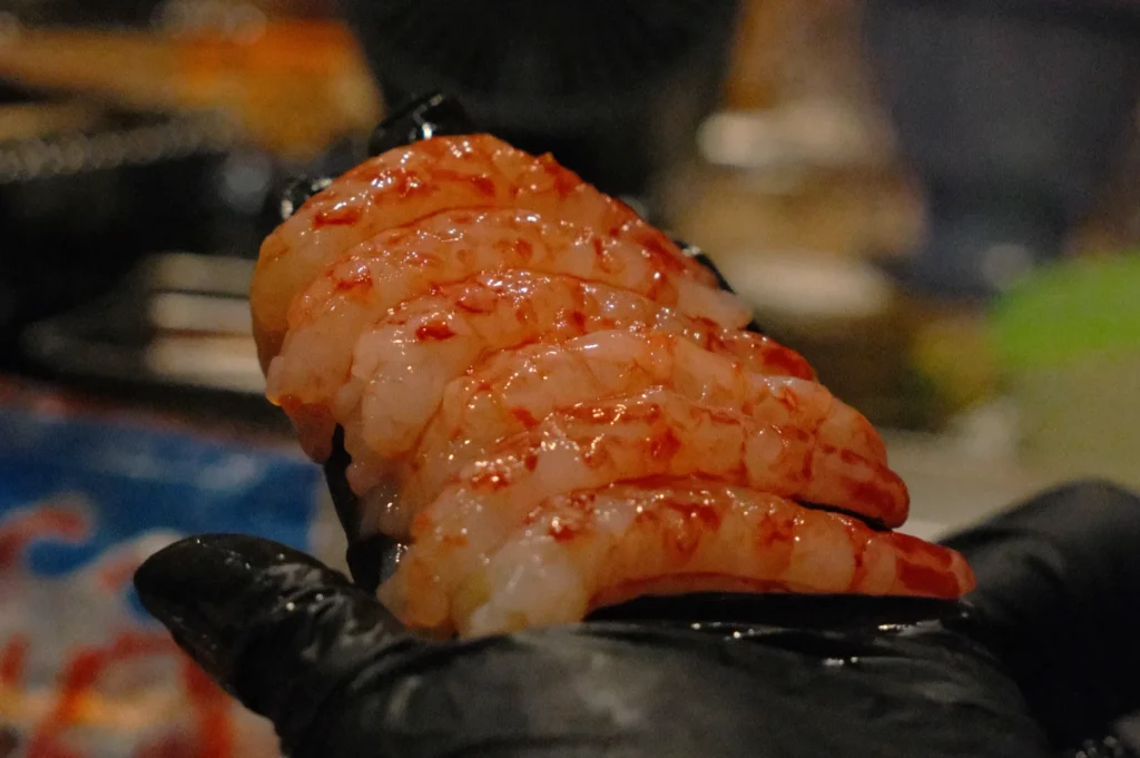 Close-up of Chef Masa’s gloved hand holding six pieces of glossy, raw shrimp nigiri sushi, contrasting against a blurred, dimly lit background.