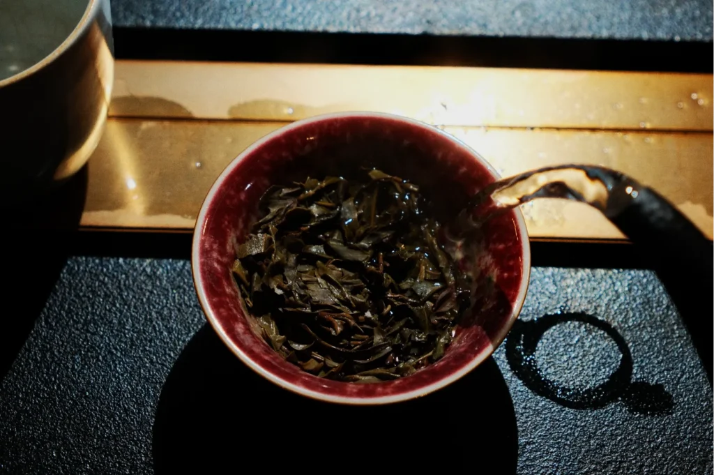 A close-up of tea leaves being steeped in a red bowl, with hot water poured from a teapot. The scene conveys warmth and serenity, with soft lighting.