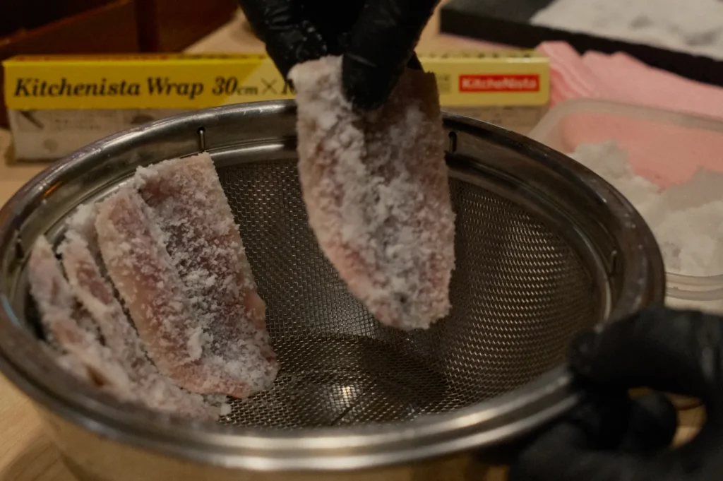 Gloved hands holding a salt-covered fish fillet over a metal strainer. Kitchen wrap and pink cloth are visible in the background.
