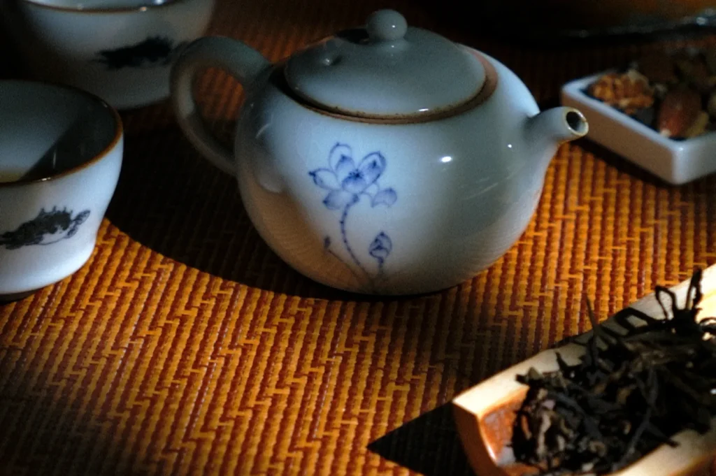 A ceramic teapot with blue floral design sits on a woven mat, surrounded by teacups and loose tea. Warm lighting creates a cozy, serene atmosphere.