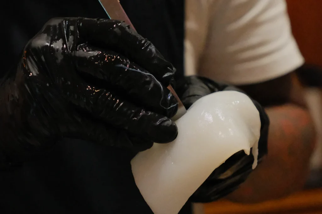 Close-up of Chef Masa’s hands in black gloves holding a knife and carving a block of white wax. The setting is likely a workshop, conveying focus and craftsmanship.