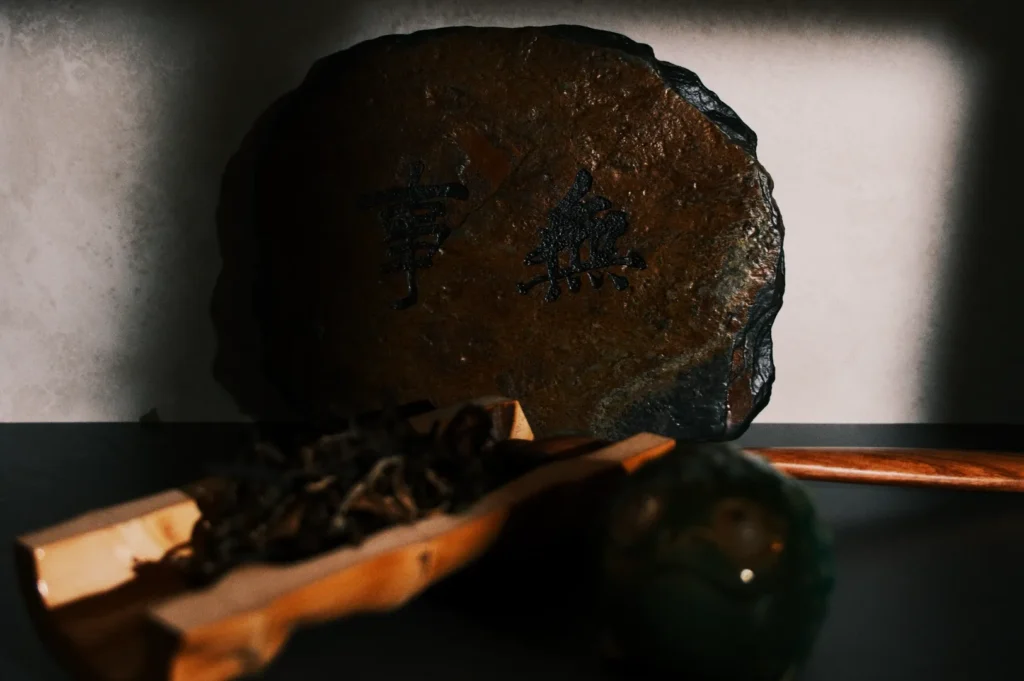 A rustic stone engraved with Asian characters stands on a table, accompanied by a wooden dish of tea leaves and a green ceramic frog, creating a serene ambiance.