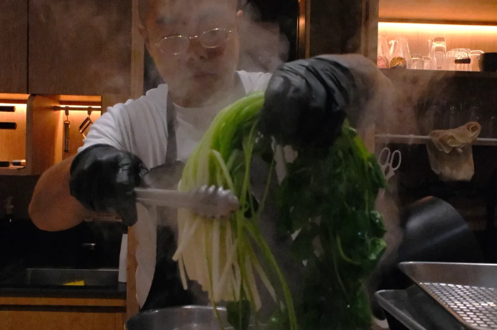 A chef wearing glasses and black gloves uses tongs to blanch leafy greens in a steaming pot. The kitchen setting has warm lighting, creating a focused and professional atmosphere.