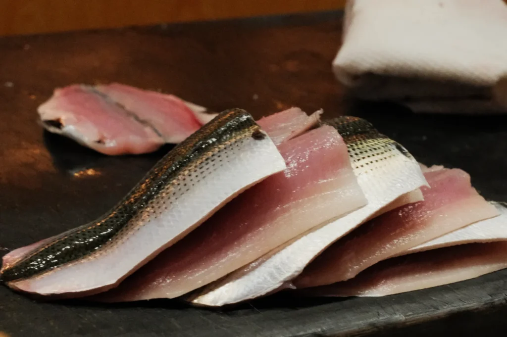 Slices of raw fish with shiny, silvery skin and pink flesh are neatly stacked on a dark cutting board. A white cloth is visible in the background.