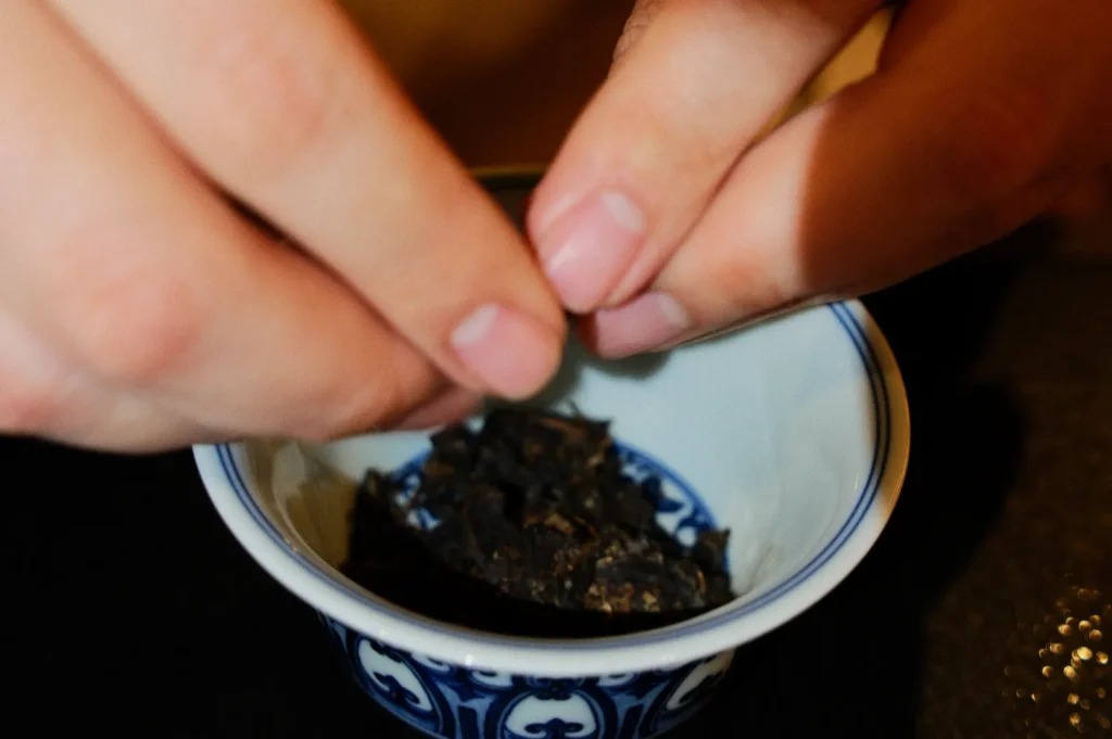 Hands gently crumble dark tea leaves into an ornate blue and white ceramic bowl, suggesting a calm and focused tea preparation ritual.