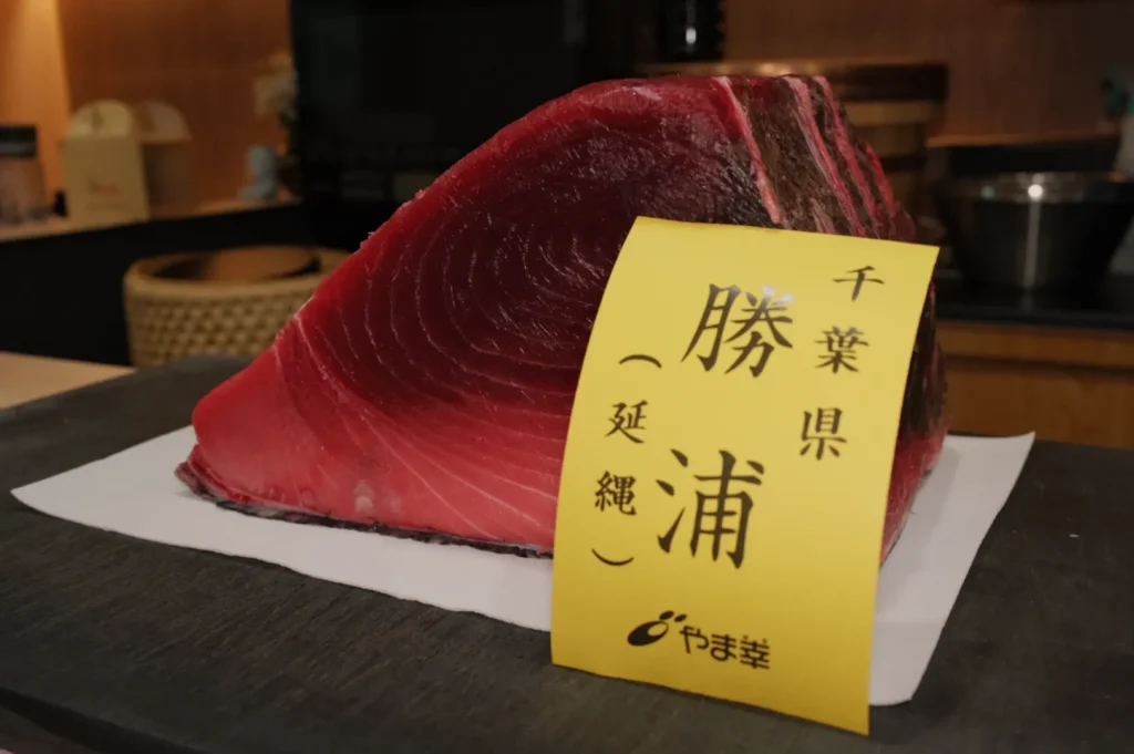 A thick, fresh cut of tuna is displayed on a white sheet with a yellow label featuring Japanese text. The scene is set in a kitchen, conveying freshness.