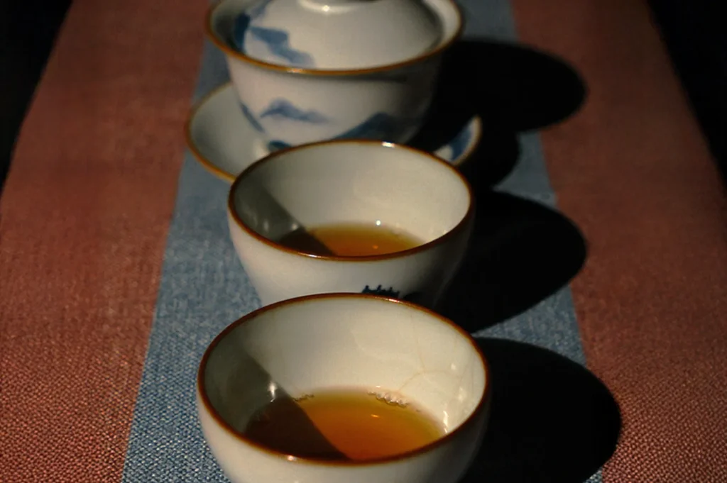 Three ceramic cups with tea and a teapot are aligned diagonally on a textured fabric. The lighting is soft, creating a warm, serene atmosphere.