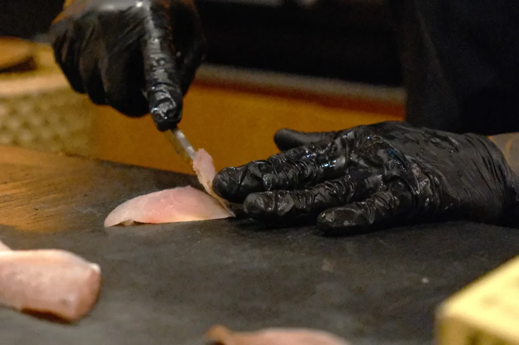 Chef Masa in black gloves skillfully slices raw fish on a dark cutting board, highlighting precision and focus in food preparation.