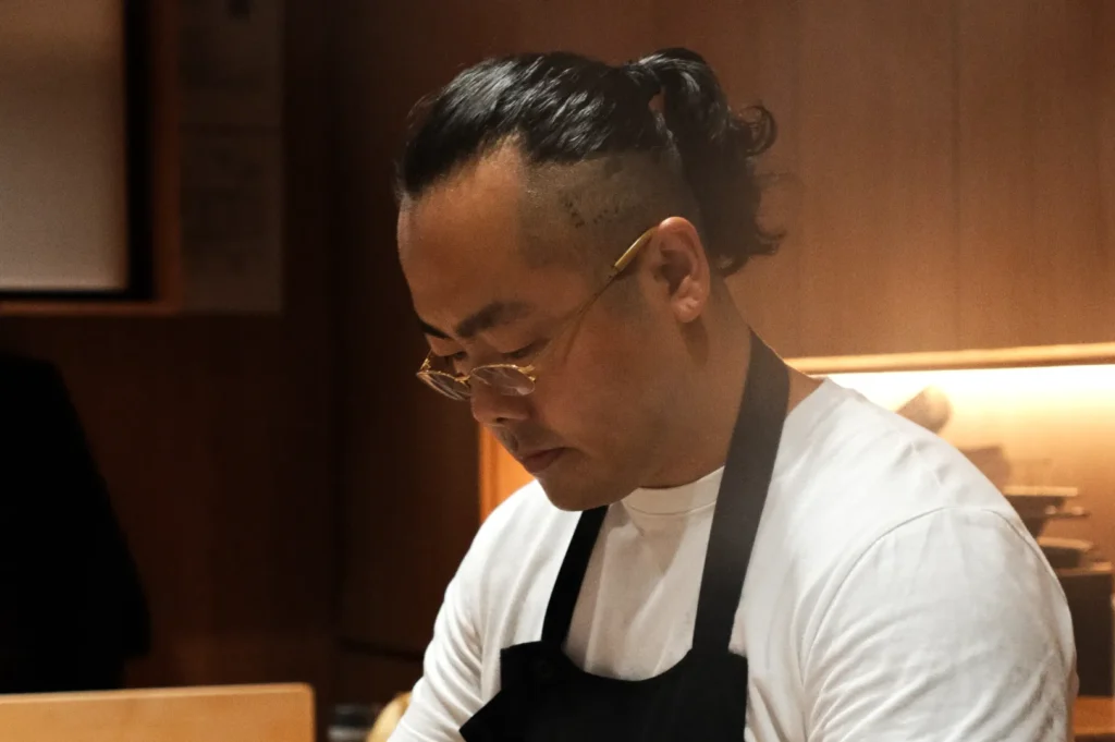 Chef Masa with a focused expression works in a warmly lit kitchen. He wears a white shirt, black apron, and glasses. The scene conveys concentration.