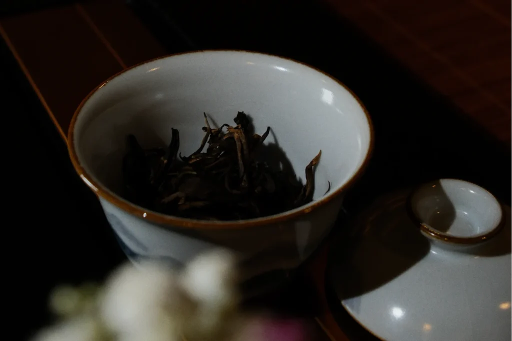 A white ceramic bowl holds dried tea leaves, placed on a dark tray. The lid rests beside it, creating a serene and elegant tea-setting atmosphere.