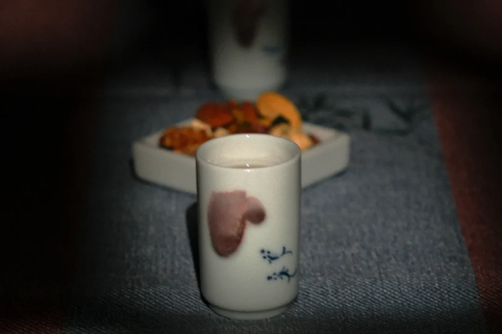 A ceramic cup with delicate floral patterns is foregrounded, set on a dark tablecloth. A blurry dish of assorted snacks sits in the background.