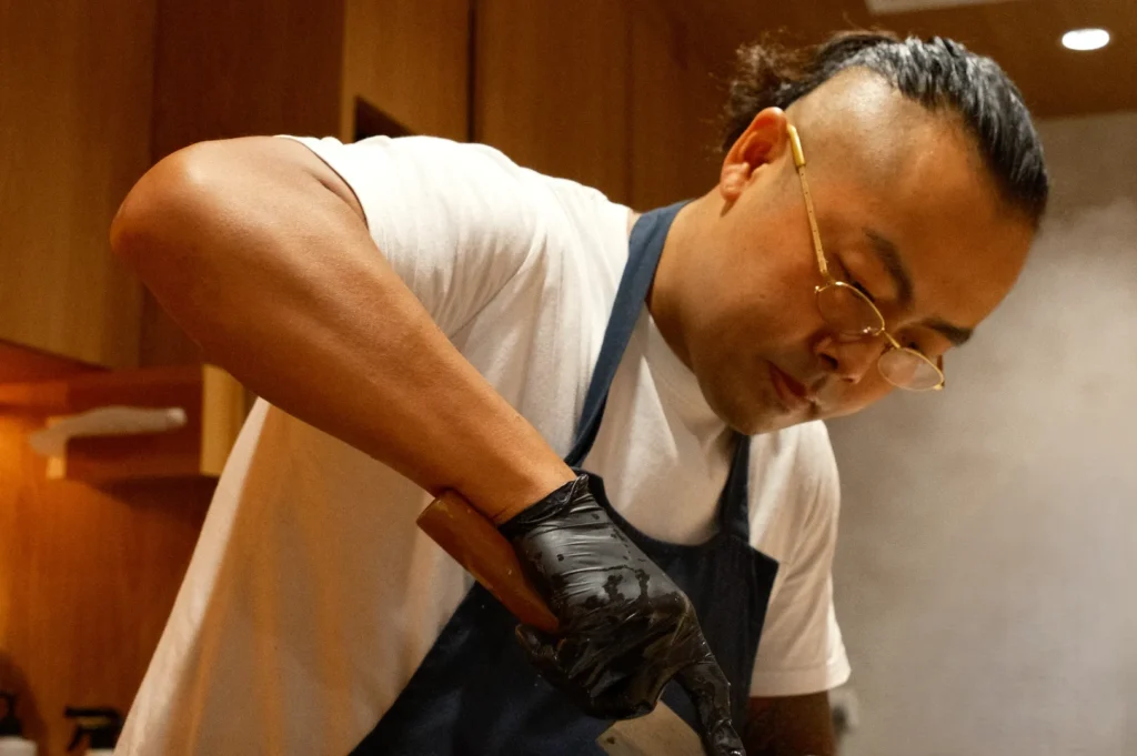 A focused Chef Masa wearing a white shirt, blue apron, and black gloves slices food with precision in a warm, wooden kitchen. Another person works in the background.