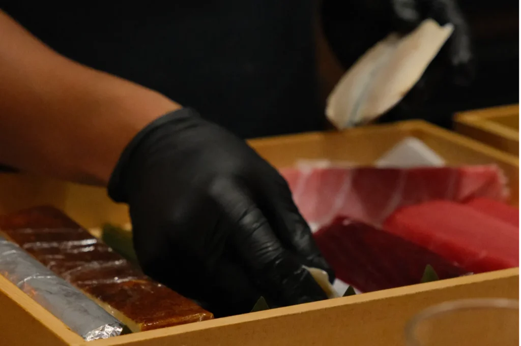 A hand in a black glove selects a piece of raw fish from a wooden tray containing an assortment of colorful sushi, conveying a focus on fresh ingredients.