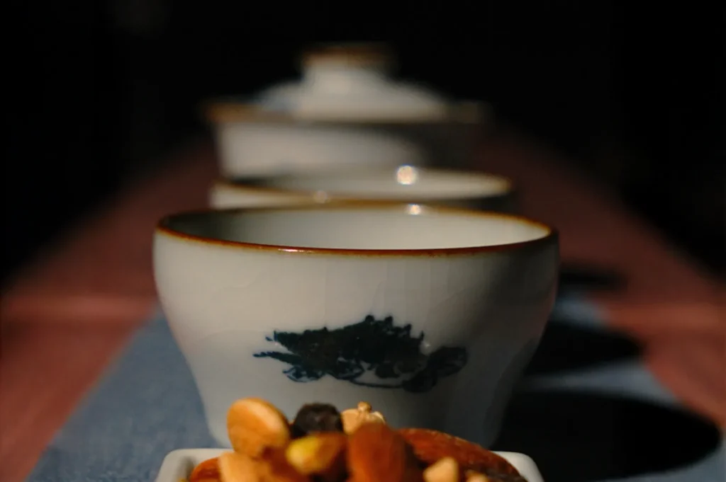 A row of white ceramic cups with a blue design, lined up on a dark table. The foreground features a dish of mixed nuts. The mood is calm and minimalistic.