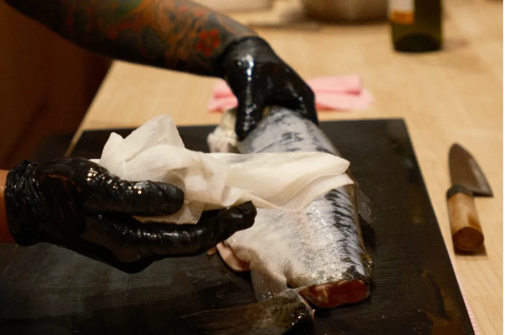 Chef Masa with a tattooed arm and black gloves cleans a large fish with a paper towel on a wooden cutting board. A knife is nearby, evoking precision and focus in food preparation.
