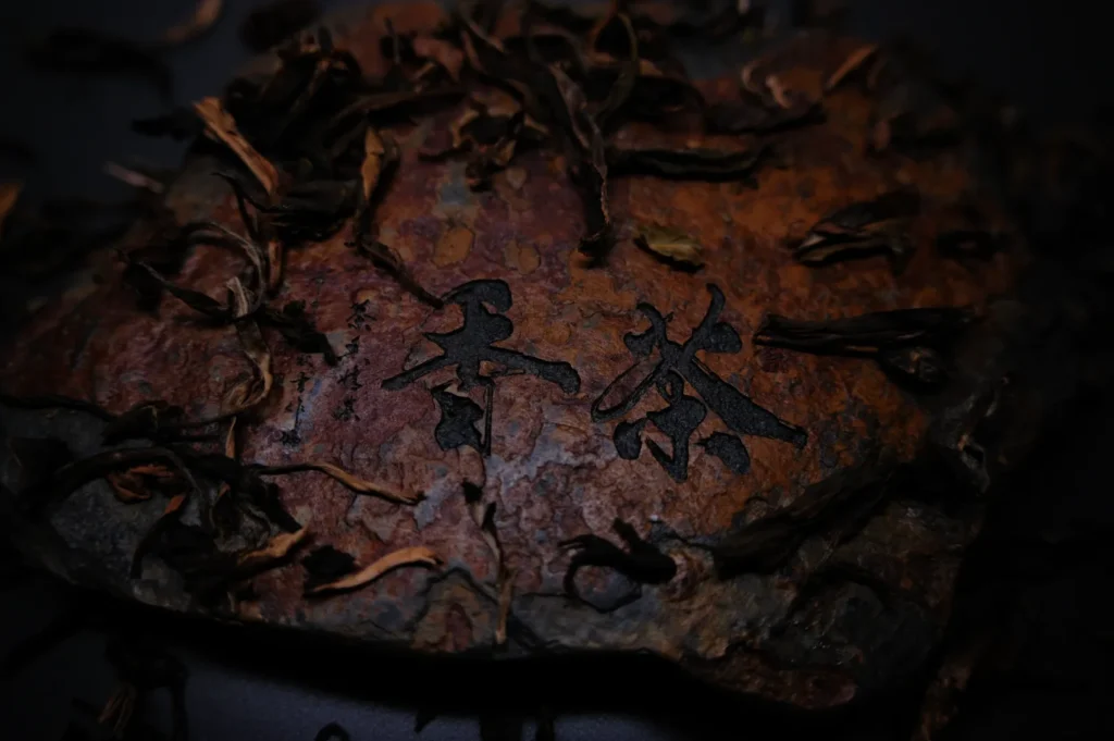 Close-up of a weathered, dried tea leaf block with dark Chinese characters on its surface. Loose tea leaves are scattered around, creating an earthy, rustic feel.