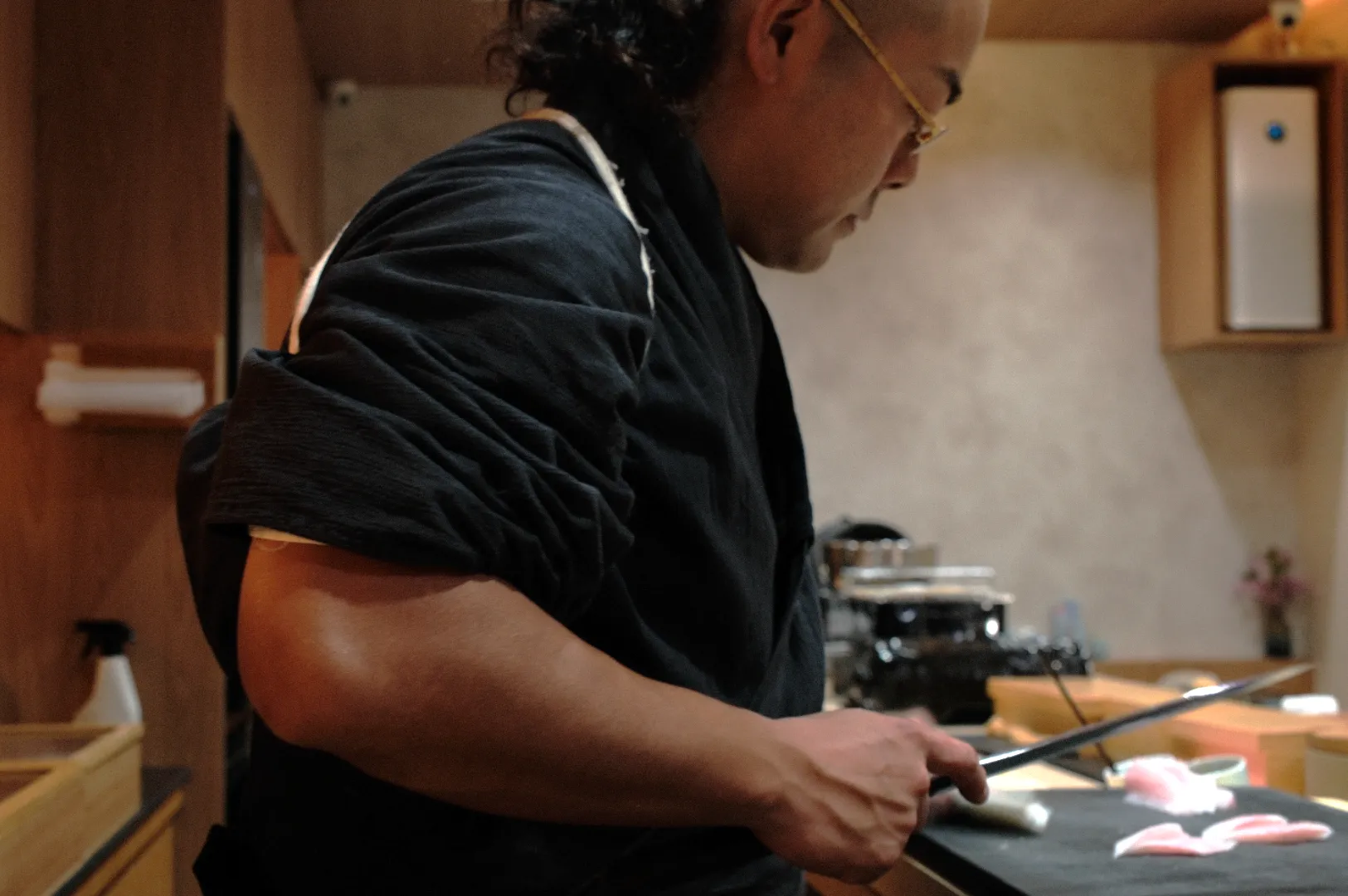 Chef Masa in black attire skillfully slices raw fish at a dimly lit sushi bar, creating a focused and tranquil ambiance.