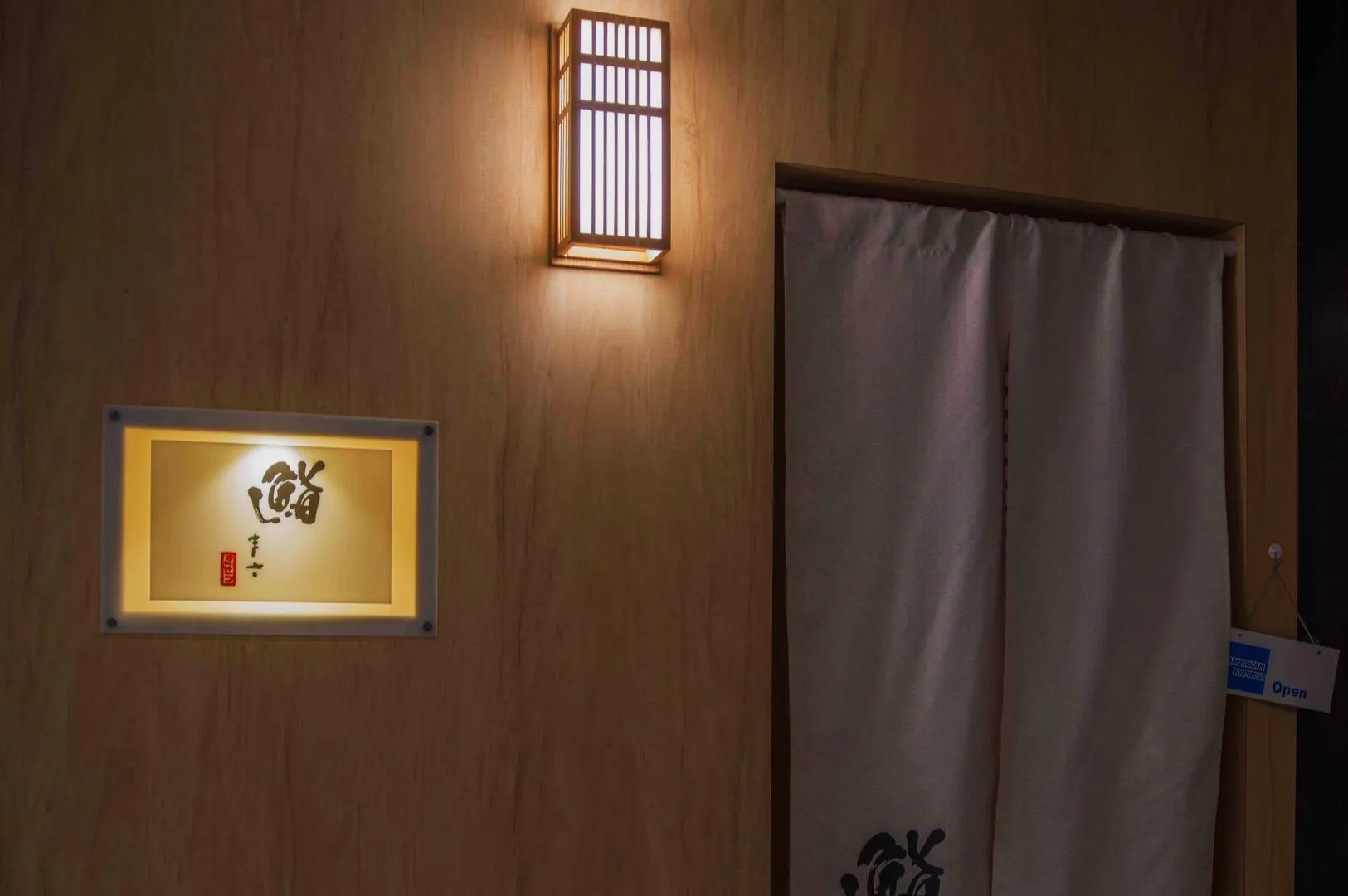 Dimly lit Japanese entrance featuring a warm wall lamp, a sign with kanji characters, and a traditional fabric curtain, creating a serene and inviting ambiance.