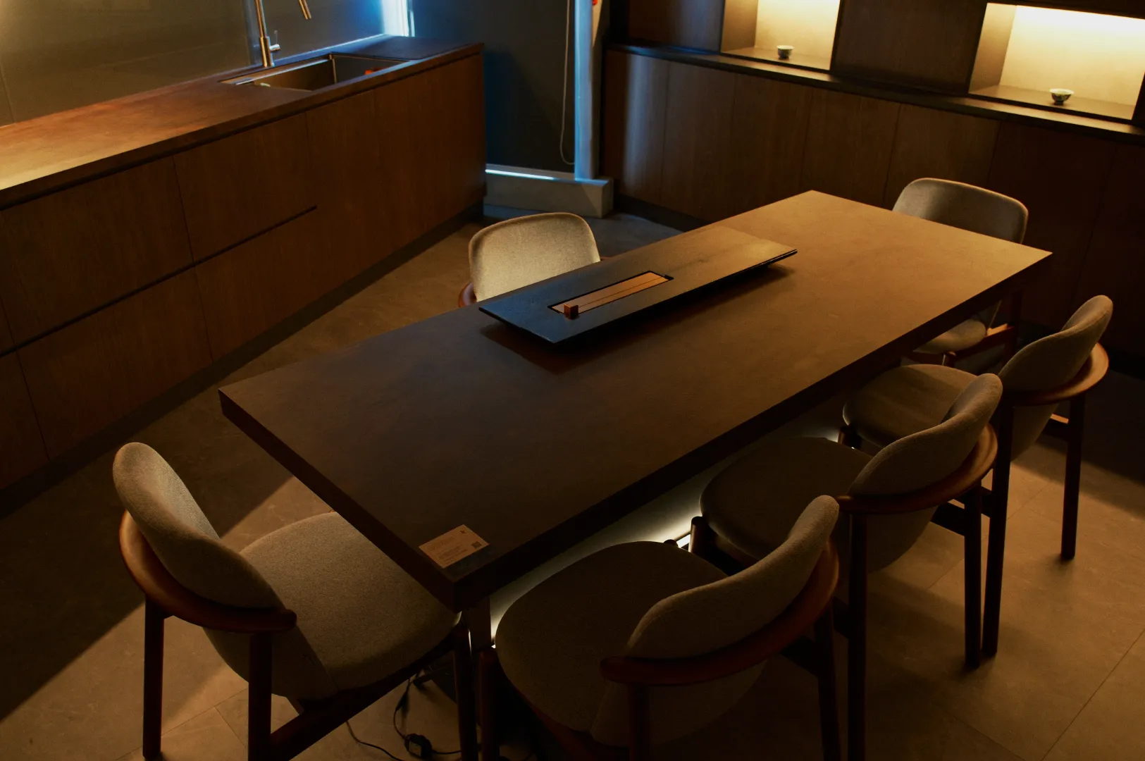 A modern kitchen with dim lighting features a long wooden table surrounded by six cushioned chairs. The sleek design creates a warm, minimalist ambiance.