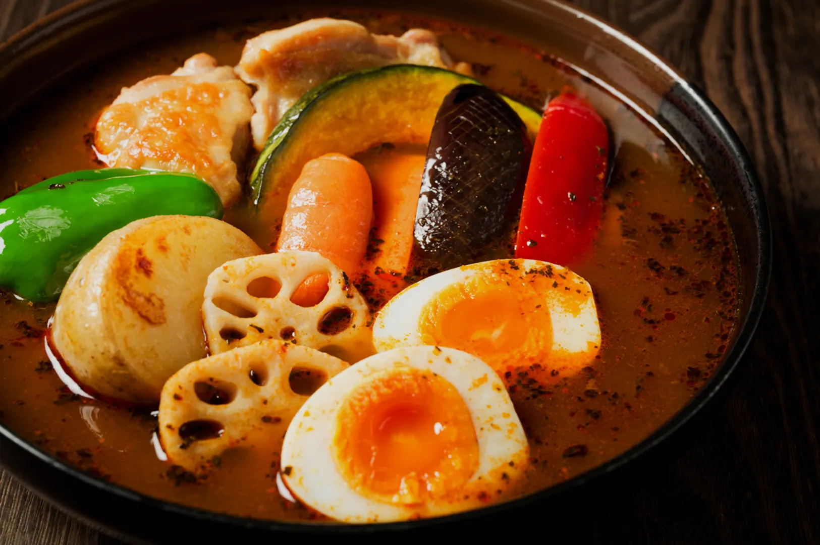 A vibrant bowl of Japanese curry soup with colorful vegetables, soft-boiled eggs, lotus root, and chicken, conveys warmth and savory richness on a wooden table.
