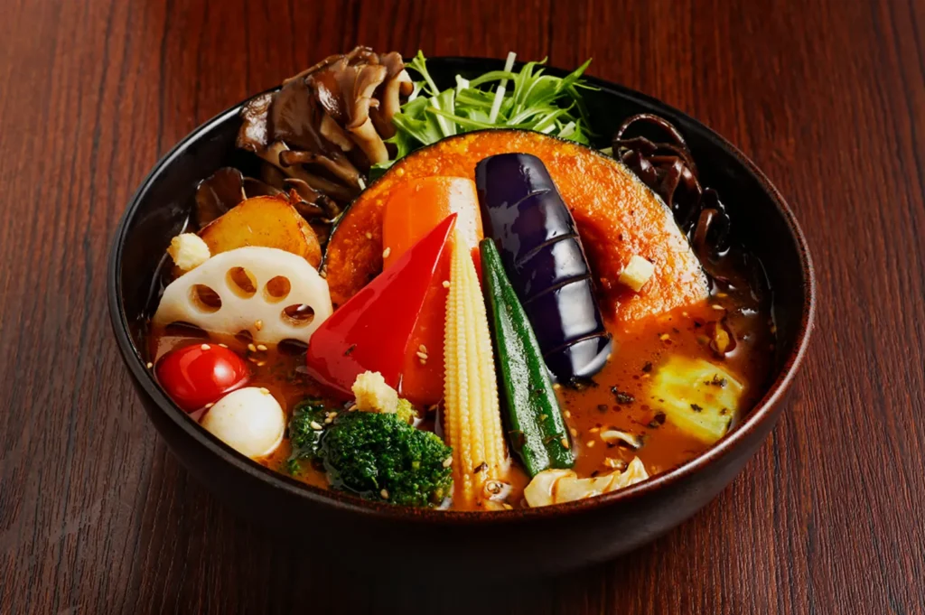 A colorful bowl of ramen on a wood table features vibrant vegetables like red pepper, eggplant, and broccoli, creating a fresh, appetizing look.