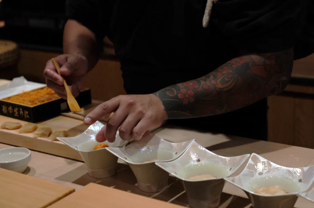 A tattooed Chef Masa carefully arranges dishes on a wooden counter, preparing food with a small utensil. The scene feels meticulous and focused.