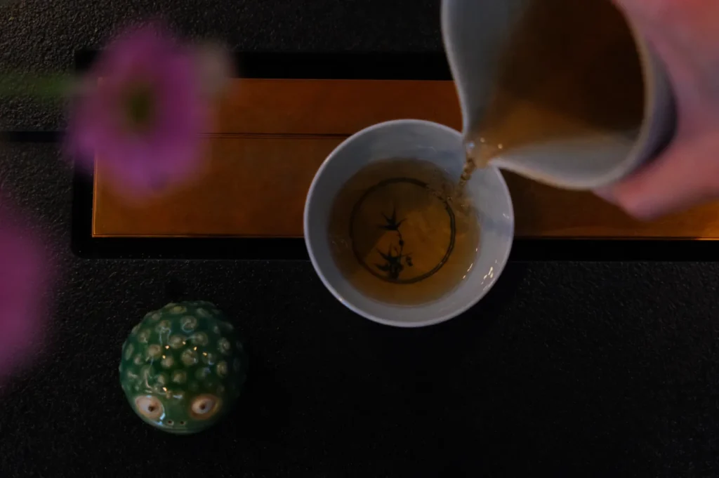 A hand pours tea into a white cup with decorative bamboo art. A small, green, textured figure and blurred pink flowers create a serene atmosphere.